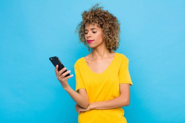 Genç Afro-Amerikalı mutlu, kendinden emin bir ifadeyle gülümsüyor, elleri çenesinde, merak ediyor ve cep telefonuyla yan tarafa bakıyor.