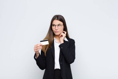 Genç ve güzel bir kadın mutlu bir şekilde gülümsüyor ve hayal kuruyor ya da şüphe ediyor, kredi kartıyla yan tarafa bakıyor.