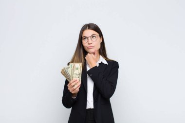 Genç, güzel bir kadın düşünüyor, şüpheci ve şaşkın hissediyor, farklı seçeneklerle, banknotlarla hangi kararı vereceğini merak ediyor.