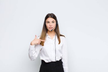 Genç ve güzel bir kadın ağzı açık, kulaklıklar ve mikrofonla kendini işaret ederek şok olmuş ve şaşırmış görünüyor.