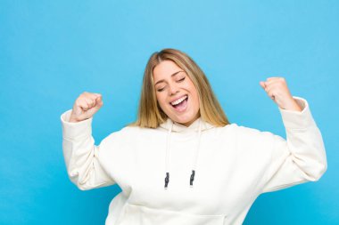 Genç sarışın kadın çok mutlu ve şaşırmış görünüyor, başarıyı kutluyor, bağırıyor ve düz duvara zıplıyor.