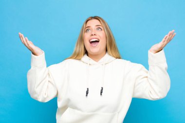 Genç sarışın kadın kendini mutlu, şaşkın, şanslı ve şaşırmış hissediyor, zaferi kutluyor, iki elini de havaya kaldırıp düz duvara yaslanıyor.