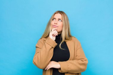 Genç sarışın kadın düşünüyor, şüpheci ve şaşkın hissediyor, farklı seçenekleri var, düz duvara karşı hangi kararı vereceğini merak ediyor.