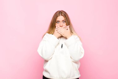 Genç sarışın kadın elleri ile ağzını kapatıyor, şok olmuş, şaşırmış bir ifadeyle, bir sır saklıyor ya da düz duvara karşı 