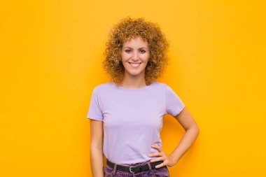 Genç afro kadın mutlu bir şekilde gülümsüyor, eli kalçasında ve kendinden emin, pozitif, gururlu ve turuncu duvara karşı dost canlısı bir tavırla.