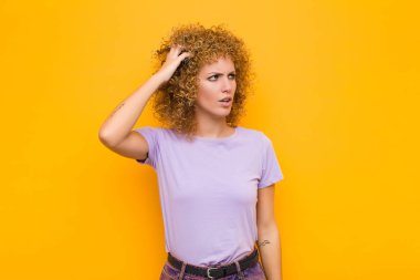 Genç afro kadın şaşkın ve şaşkın hissediyor, başını kaşıyor ve turuncu duvara bakıyor.