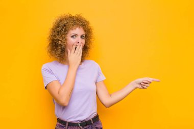 Genç afro kadın kendini mutlu, şaşkın ve şaşırmış hissediyor. Ağzı eliyle kaplıyor ve turuncu duvara karşı yanal fotokopi alanını işaret ediyor.