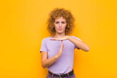 Genç afro kadın ciddi, sert, kızgın ve hoşnutsuz görünüyor, turuncu duvara karşı zaman geçirme işareti yapıyor.