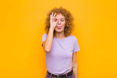 Genç afro kadın mutlu bir şekilde gülümsüyor, şaka yapıyor ve gözetleme deliğinden bakıyor, turuncu duvara karşı sırları gözetliyor.