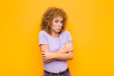 Genç afro kadın omuz silkiyor, kafası karışık ve kararsız, kolları bağlı ve şaşkın turuncu duvara bakıyor.