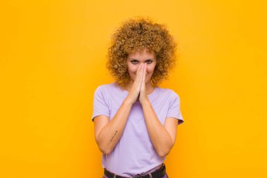 Genç afro kadın endişeli, umutlu ve dindar hissediyor, avuçlarına bastırılarak sadık bir şekilde dua ediyor, turuncu duvara karşı af diliyor.