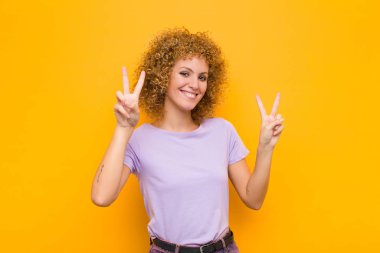 Genç afro kadın gülümsüyor ve mutlu görünüyor, arkadaş canlısı ve memnun, iki eliyle turuncu duvara karşı zafer ya da barış işareti yapıyor.