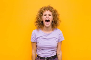 Genç afro kadın saldırganca bağırıyor, çok kızgın, sinirli, öfkeli ya da sinirli görünüyor, turuncu duvara karşı hayır diye bağırıyor.