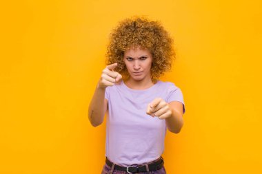 Genç afro kadın kameraya hem parmaklarını hem de kızgın yüzünü göstererek turuncu duvara karşı görevini yapmanı söylüyor.