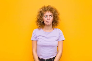 Genç afro kadın şaşkın ve şaşkın görünüyor, merak ediyor veya bir sorunu çözmeye çalışıyor veya turuncu duvara karşı düşünüyor.