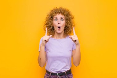 Genç afro kadın korkuyla ve ağzı açık bir şekilde turuncu duvara doğru şaşkın ve şaşkın bir bakışla yukarıya bakıyor.