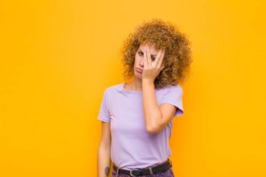 Genç afro kadın yorgun, sıkıcı ve sıkıcı bir görevden sonra sıkılmış, hayal kırıklığına uğramış ve uykulu hissediyor, yüzünü turuncu duvara dayamış.