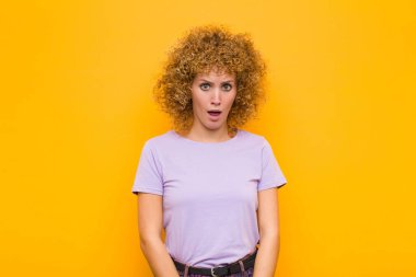 Genç afro kadın şaşkın ve şaşkın hissediyor, aptal, afallamış bir ifadeyle turuncu duvara karşı beklenmedik bir şeye bakıyor.