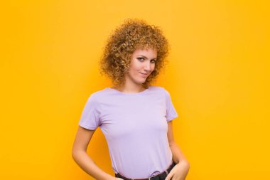 Genç afro kadın gururlu, kendinden emin, havalı, küstah ve küstah görünüyor, gülümsüyor, turuncu duvara karşı başarılı hissediyor.