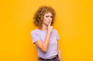 Genç afro kadın sessizlik ve sessizlik istiyor, parmağıyla ağzının önünde işaret ediyor, ya da turuncu duvara karşı bir sır saklıyor.