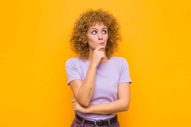 Genç afro kadın düşünüyor, kuşkulu ve şaşkın hissediyor, farklı seçeneklerle, turuncu duvara karşı hangi kararı vereceğini merak ediyor.