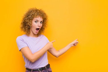Genç afro kadın şok olmuş ve şaşırmış hissediyor, yan taraftaki fotokopi alanını işaret ederek, turuncu duvara karşı şaşkın, açık ağızlı bir bakışla