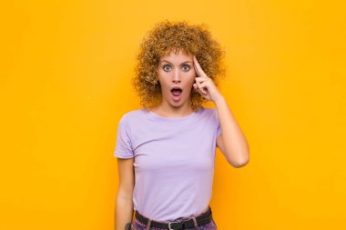 Genç afro kadın şaşırmış, ağzı açık, şok olmuş görünüyor, turuncu duvara karşı yeni bir düşünce, fikir ya da konsept fark ediyor.