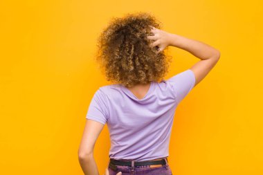 Genç afro kadın bilgisiz ve kafası karışık, bir çözüm düşünüyor, eli kalçasında, diğeri kafasında, turuncu duvara karşı dikiz manzaralı