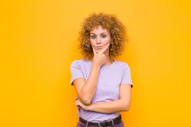Genç afro kadın ciddi, düşünceli ve güvensiz görünüyor, bir kolu çapraz ve çene üzerinde, turuncu duvara karşı seçenekleri tartıyor