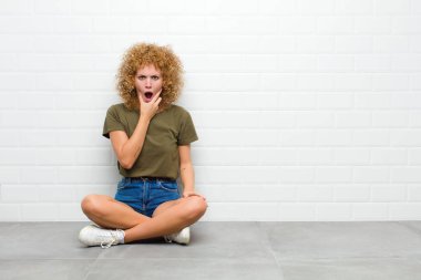 Ağzı ve gözleri açık genç afro bir kadın ve elleri çenesinde, hoş olmayan bir şekilde şokta, yerde oturmuş ne ya da wow diyor.