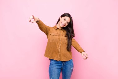 Genç ve güzel bir kadın mutlu ve kendinden emin, kamerayı iki eliyle gösteriyor ve gülüyor, pembe duvara karşı seni seçiyor.