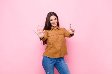 Genç, güzel bir kadın gülümsüyor ve arkadaş canlısı görünüyor. Elleri önde, pembe duvara doğru geri sayarak yedi ya da yedinci numarayı gösteriyor.