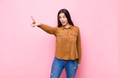 Genç, güzel bir kadın şaşkın ve şaşırmış hissediyor. Pembe duvara karşı hayret dolu, açık ağızlı bir bakışla yukarılara bakıyor.