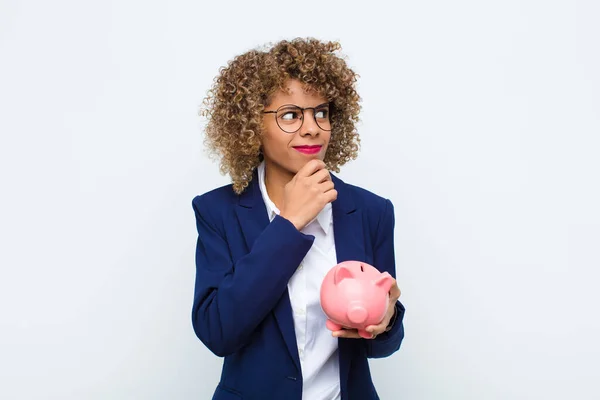 young african american woman thinking, feeling doubtful and confused, with different options, wondering which decision to make with a piggy bank