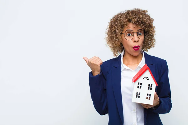 young african american woman looking astonished in disbelief, pointing at object on the side and saying wow, unbelievable with house model