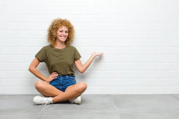 young afro woman feeling happy and cheerful, smiling and welcoming you, inviting you in with a friendly gesture sitting on a floor