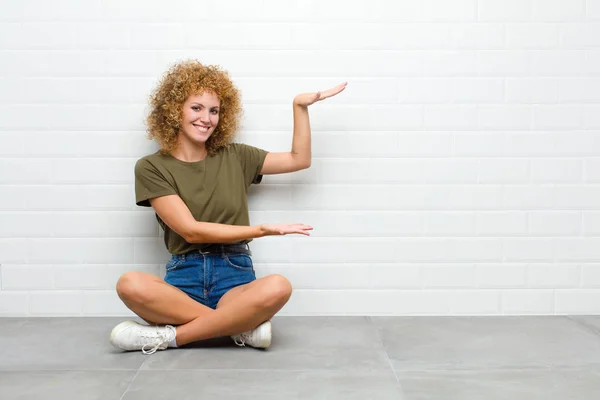young afro woman smiling, feeling happy, positive and satisfied, holding or showing object or concept on copy space sitting on a floor