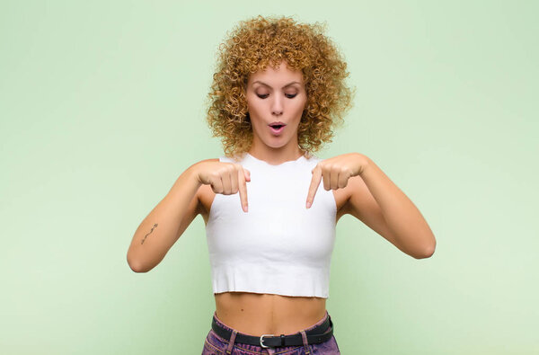 young afro woman with open mouth pointing downwards with both hands, looking shocked, amazed and surprised against green wall