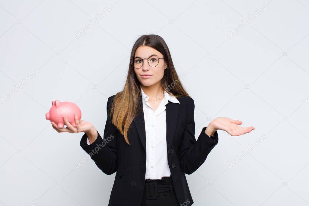 young pretty woman feeling puzzled and confused, doubting, weighting or choosing different options with funny expression with a piggy bank