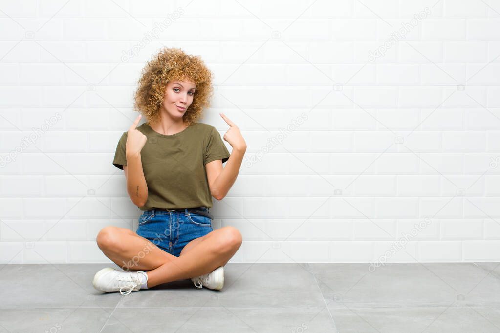 young afro woman with a bad attitude looking proud and aggressive, pointing upwards or making fun sign with hands sitting on a floor