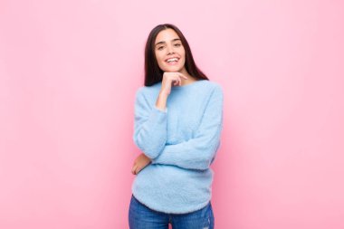 Genç ve güzel bir kadın mutlu görünüyor ve gülümsüyor, elleri çenesinde, merak ediyor ya da soru soruyor, pembe duvarla seçenekleri karşılaştırıyor.