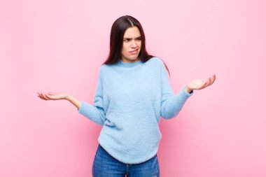 Genç, güzel bir kadın şaşkın, kafası karışmış ve stresli görünüyor, farklı seçenekler arasında merak ediyor, pembe duvara karşı kararsız hissediyor.