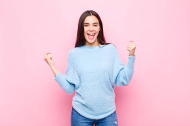 Genç ve güzel bir kadın son derece mutlu ve şaşırmış görünüyor, başarıyı kutluyor, bağırıyor ve pembe duvara zıplıyor.