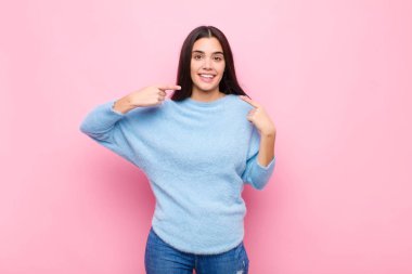 Genç, güzel bir kadın, kendine güvenerek gülümseyerek pembe duvara karşı pozitif, rahat ve tatmin edici bir tavır sergiliyor.
