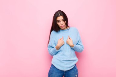 Genç ve güzel bir kadın, şaşkın ve tuhaf bakışlarıyla kendini işaret ediyor. Pembe duvara karşı seçildiği için şaşırmış ve şaşırmış.