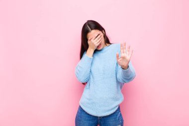 Genç ve güzel bir kadın yüzünü elleriyle kapatıyor ve kamerayı durdurmak için diğer elini öne koyuyor. Pembe duvara karşı fotoğraf ya da resim çekmeyi reddediyor.