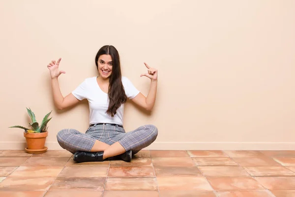 young pretty woman framing or outlining own smile with both hands, looking positive and happy, wellness concept sitting on a floor