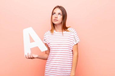 Genç, güzel, kızıl saçlı kadın, kafası karışmış, şüpheli, düşünüyor, bir kelime ya da cümle oluşturmak için alfabenin A harfini tutuyor..