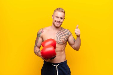 Genç sarışın güçlü adam kendini mutlu, şaşkın, tatmin olmuş ve şaşırmış hissediyor, iyi olduğunu gösteriyor ve el kol hareketleri yapıyor, boks eldivenleriyle gülümsüyor.