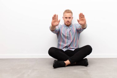 Genç sarışın adam ciddi, mutsuz, kızgın ve hoşnutsuz görünüyor. Ya da her iki avuç içi çimento zemininde otururken dur diyor.